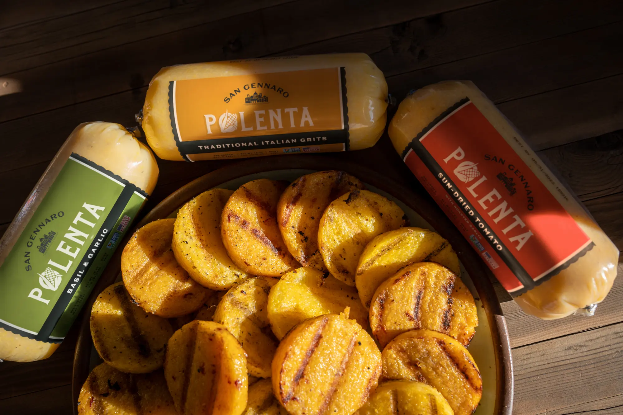 A plate of grilled polenta rounds on a wood table, ringed by the three rolls: Basil & Garlic, Traditional Italian and Sun-Dried Tomato.